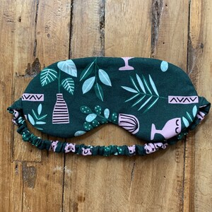 May include: A dark green sleep mask with a botanical pattern featuring pink and white plant illustrations. The mask has an elastic band with a matching floral design. The sleep mask is designed to block out light for better sleep.