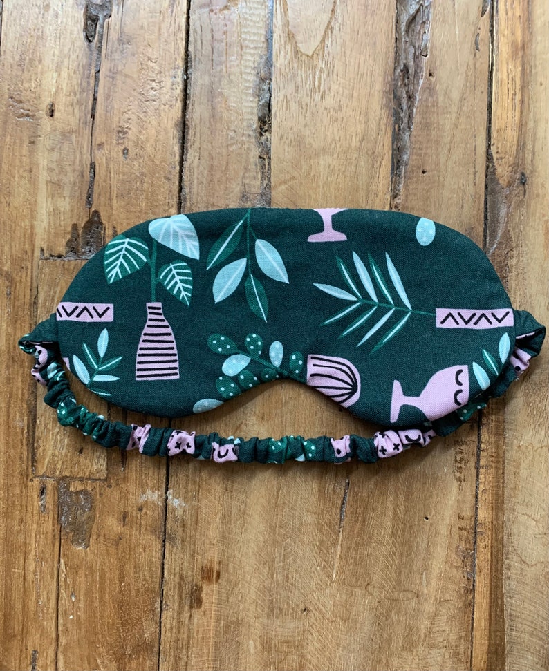 May include: A dark green sleep mask with a botanical pattern featuring pink and white plant illustrations. The mask has an elastic band with a matching floral design. The sleep mask is designed to block out light for better sleep.