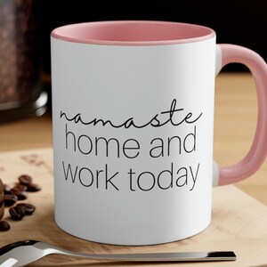Puede incluir: Taza de café de cerámica blanca con borde y asa de color rosa. La taza tiene texto negro que dice "namaste home and work today".