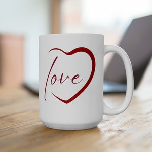 Puede incluir: Taza de café de cerámica blanca con un corazón rojo y la palabra "love" escrita dentro del corazón.