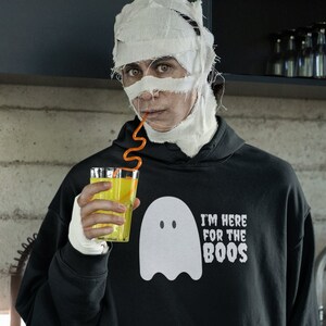 May include: A black hoodie with a white ghost graphic and the text "I'M HERE FOR THE BOOS." The person in the image is wearing a mummy costume and drinking from a glass with a yellow liquid.