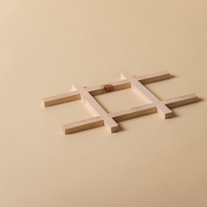 May include: A wooden tic-tac-toe game board made of light-colored wood. The game board is constructed from rectangular wooden pieces arranged in a grid pattern. The background is a solid, light tan color.