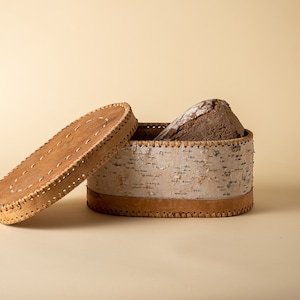 Puede incluir: Una caja de pan redonda y ovalada hecha de corteza de abedul con una tapa de cuero. La tapa está abierta y hay una hogaza de pan dentro de la caja.