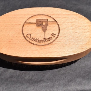 May include: Oval-shaped wooden brush with a handle cutout. The light-colored wood has a laser-etched design in a circle that says "Customize It" with a graphic. The brush has bristles on the bottom.