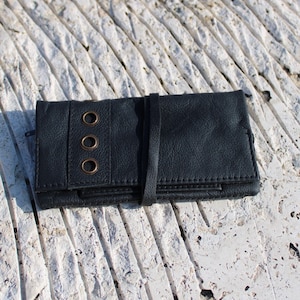 Peut inclure: Un portefeuille en cuir noir avec une fermeture à sangle et trois œillets en laiton. Le portefeuille est posé sur une surface texturée blanche.