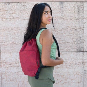 Może przedstawiać: Kobieta w zielonej koszulce na ramiączkach i czerwonym plecaku ze sznurkiem. Plecak ma czarny sznurek i czarny pasek.