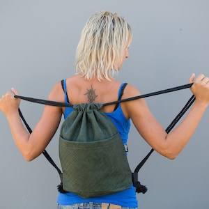 Op de afbeelding: Een vrouw in een blauwe tanktop houdt een groene rugzak met trekkoord en zwarte banden vast. De rugzak heeft een ritsvak aan de voorkant.