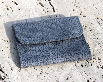 May include: A small, blue wallet with a textured surface and a flap closure. The wallet is rectangular and appears to be made of leather or a similar material. The color is a muted blue-gray, and the texture gives it a unique appearance.