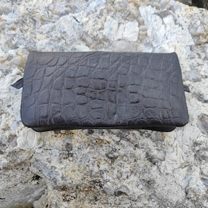 Könnte beinhalten: Eine rechteckige, dunkelbraune Tasche mit einer strukturierten, geprägten Krokodilleder-Optik. Das Accessoire hat eine glatte, flache Oberseite und eine strukturierte Form mit kleinen seitlichen Laschen an den linken und rechten Rändern.