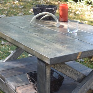 May include: A rustic wooden coffee table with a gray stained finish. The table has a lower shelf and a criss-cross design on the legs. The table is decorated with antlers, a glass, and a basket.