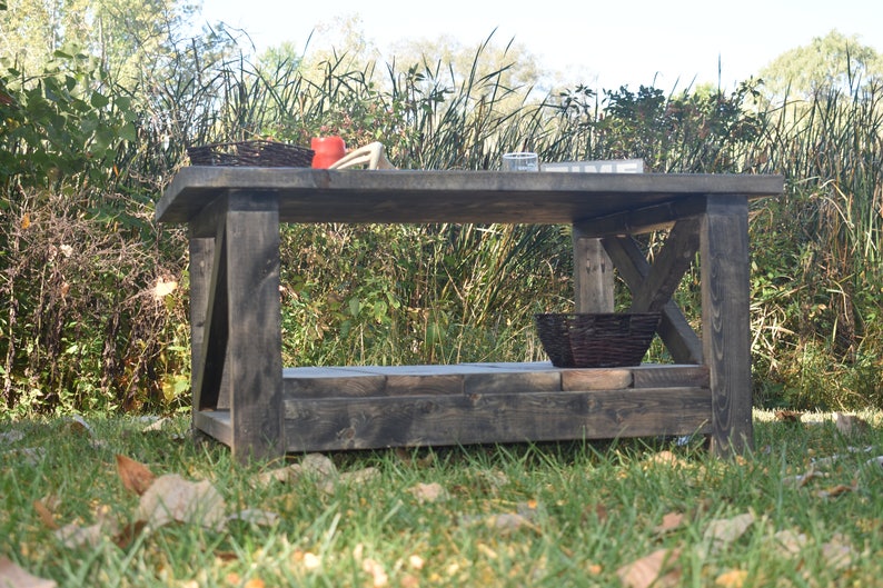 May include: A dark gray wooden coffee table with a lower shelf. The table has a rustic, farmhouse style and is made of reclaimed wood. The table is sitting on a grassy lawn with fallen leaves.