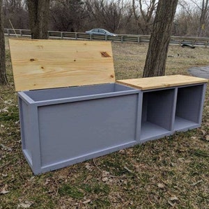 May include: A gray wooden storage bench with a hinged lid and two open cubbies. The lid and top of the cubbies are natural wood. The bench is outdoors on grass.