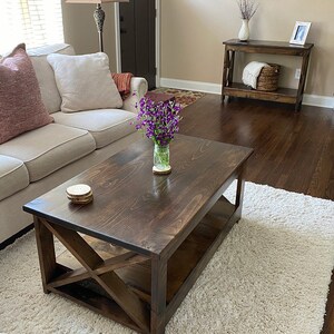 May include: A dark brown wooden coffee table with a lower shelf and an X-shaped design on the legs. The table is sitting on a white shag rug in a living room setting.