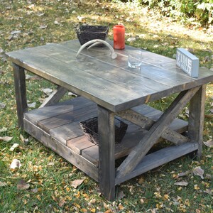May include: A rustic wooden coffee table with a lower shelf. The table is stained a dark gray color and has a distressed finish. The table is sitting on a grassy lawn with fallen leaves.