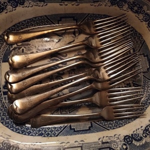 May include: A set of antique silver forks displayed on a blue and white patterned oval serving dish. The forks show signs of tarnish, and each has an engraved initial on the handle. The dish has a classic, vintage aesthetic.