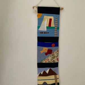 May include: A handmade textile wall hanging with an abstract nautical design. The artwork features a sailboat, a lighthouse, and a mountain scene, with a color palette of blue, black, red, and yellow. The piece is suspended from a wooden dowel.