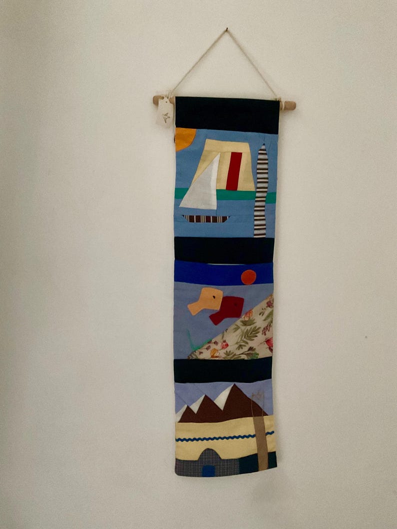 May include: A handmade textile wall hanging with an abstract nautical design. The artwork features a sailboat, a lighthouse, and a mountain scene, with a color palette of blue, black, red, and yellow. The piece is suspended from a wooden dowel.