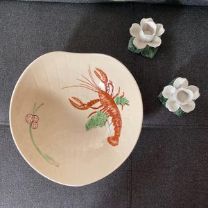 Puede incluir: Un cuenco de cerámica color crema con una ilustración de langosta roja y detalles verdes. También hay dos candelabros blancos con forma de flor y hojas verdes. El cuenco tiene una forma única.