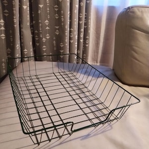 May include: A green wire desk organiser tray. The tray is rectangular with a grid pattern and slightly raised sides. The metal has a dark green finish. The tray is empty and sits on a white surface.