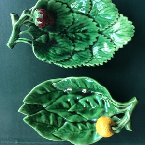 May include: Two ceramic leaf-shaped dishes. One has a red strawberry accent, the other a yellow lemon. The dishes are a glossy green with detailed leaf veining. Ideal for serving small portions or as decorative pieces.