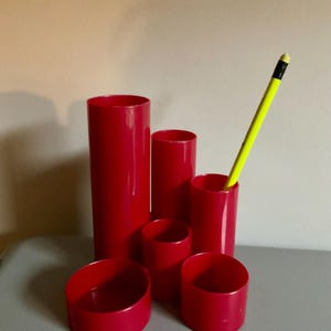 May include: A set of six red cylindrical containers of varying heights arranged in a cluster, with a bright yellow pencil placed inside the tallest of the shorter cylinders. The containers have smooth, solid red surfaces and open tops.