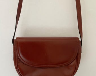 May include: A brown leather crossbody bag with a curved flap and a long, thin strap. The bag has a glossy finish and a simple, classic design. The strap is attached to the bag with small metal hardware.