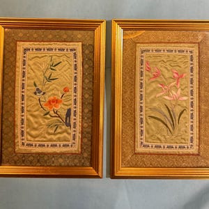 May include: A pair of framed silk embroidered panels. One displays a floral design with orange and blue details, the other features pink flowers. Both are in gold frames with decorative borders, offering a traditional look.