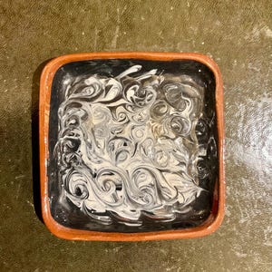 May include: Square ceramic dish with a black interior and a swirling white and black pattern. The rim is a warm brown color. The dish is likely used for serving or display.