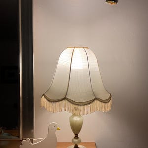 Peut inclure: Une lampe de table de style vintage avec un abat-jour crème et une bordure à franges. La lampe a un socle décoratif et se trouve à côté d'une figurine d'oiseau en bois blanc. Deux décorations murales d'oiseaux sont au-dessus de la lampe.