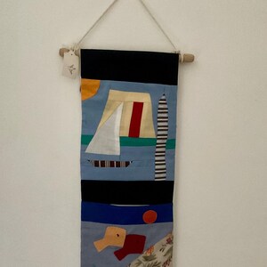 May include: A colorful, abstract wall hanging with a nautical theme. The textile art piece features a sailboat, a striped tower, and geometric shapes in shades of blue, black, red, and yellow. It is suspended from a wooden dowel with a natural cord.