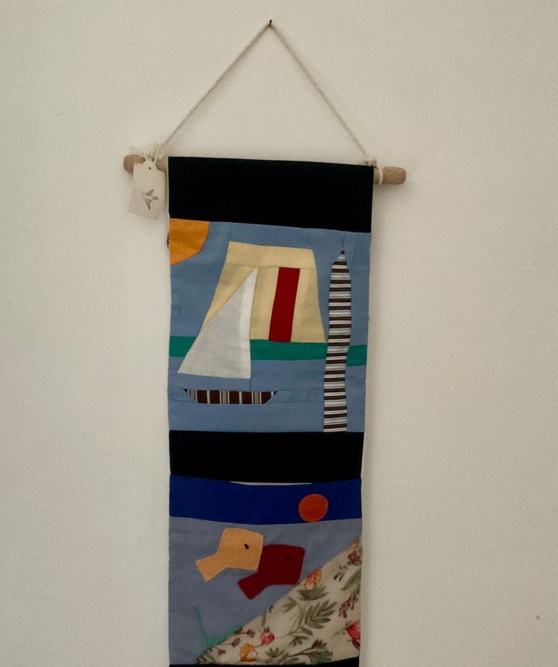 May include: A colorful, abstract wall hanging with a nautical theme. The textile art piece features a sailboat, a striped tower, and geometric shapes in shades of blue, black, red, and yellow. It is suspended from a wooden dowel with a natural cord.