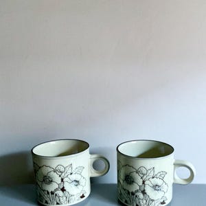May include: Two cream ceramic mugs with a floral design. The mugs have a dark brown rim and handle. The floral design features white flowers with brown outlines and green leaves. The mugs are placed on a light blue surface.