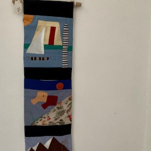 May include: A vertical textile wall hanging with abstract geometric designs. The top section is black, followed by blue, with a sailboat, a red stripe, and a striped tower. Other sections include a red circle, shapes, and a mountain scene.