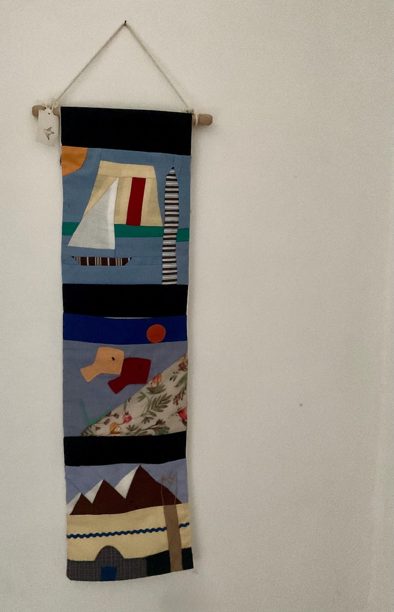 May include: A vertical textile wall hanging with abstract geometric designs. The top section is black, followed by blue, with a sailboat, a red stripe, and a striped tower. Other sections include a red circle, shapes, and a mountain scene.