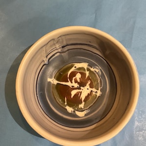 Könnte beinhalten: Eine handgefertigte Keramikschale mit cremefarbenem Rand und blau-braunem Innenraum. Die Mitte der Schale zeigt ein braunes und grünes Design mit weißen Akzenten, wodurch ein einzigartiger, künstlerischer Look entsteht. Ideal zum Servieren von Suppen, Salaten oder anderen Gerichten.