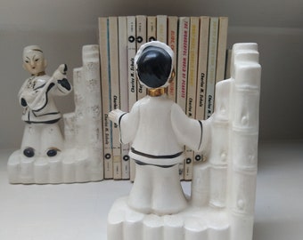 May include: A pair of vintage ceramic bookends. One bookend depicts a figure playing a stringed instrument, the other a figure with arms outstretched. Both figures are dressed in white robes with black accents. A row of books is visible between the bookends.