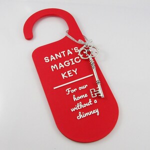 May include: A red door hanger with a curved top and the text "SANTA'S MAGIC KEY" in white. A silver key is attached with a silver ribbon. The text below reads "For our home without a chimney."