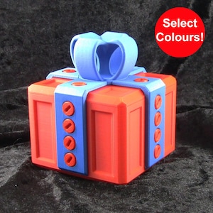 May include: A red gift box with a blue ribbon and blue and red accents. The box has a screw-on lid and is decorated with a bow on top. "Select Colours!" is written in red text on a white circle.