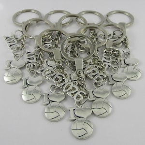 May include: A collection of silver-toned metal keychains, each featuring a split ring, a chain, the number 2026, a letter I, a heart shape, and a volleyball charm. The charms are arranged in a cluster, showcasing the metallic finish and details.