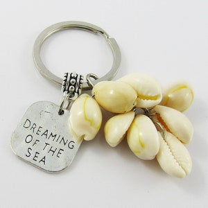 May include: A silver keyring adorned with cream-coloured seashells. A small, square metal charm reads "Dreaming of the Sea". The keyring features a silver ring and decorative bead.