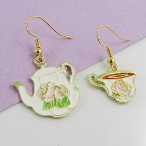 May include: A pair of gold earrings featuring a white teapot and a white teacup design. The teapot and teacup are decorated with pink roses and green leaves.