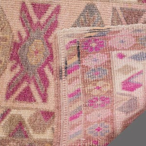 May include: Close-up of a vintage rug with a faded, distressed appearance. The rug features geometric patterns in shades of pink, beige, and gray. The design includes diamond and floral motifs, with a folded corner revealing the rug's backing.