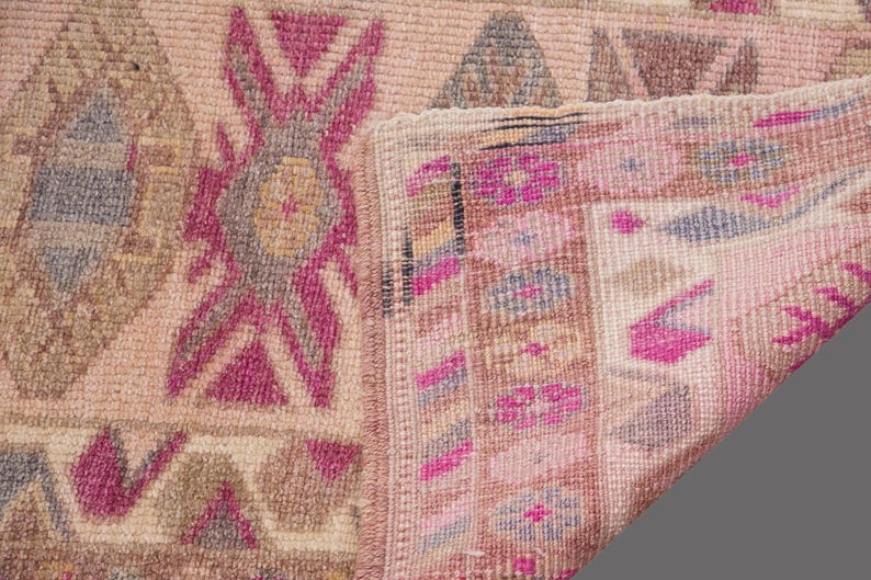 May include: Close-up of a vintage rug with a faded, distressed appearance. The rug features geometric patterns in shades of pink, beige, and gray. The design includes diamond and floral motifs, with a folded corner revealing the rug's backing.