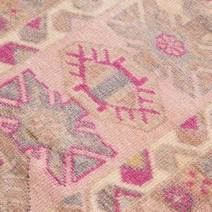 May include: Close-up of a vintage rug with a repeating geometric pattern. The rug features a soft pink background with accents of brown, gray, and fuchsia. The design includes stylized shapes and floral motifs, creating a textured appearance.