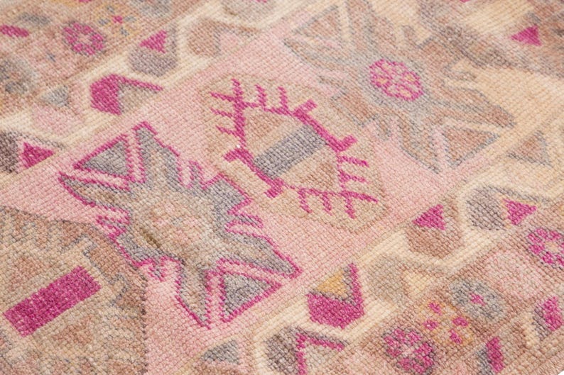 May include: Close-up of a vintage rug with a repeating geometric pattern. The rug features a soft pink background with accents of brown, gray, and fuchsia. The design includes stylized shapes and floral motifs, creating a textured appearance.