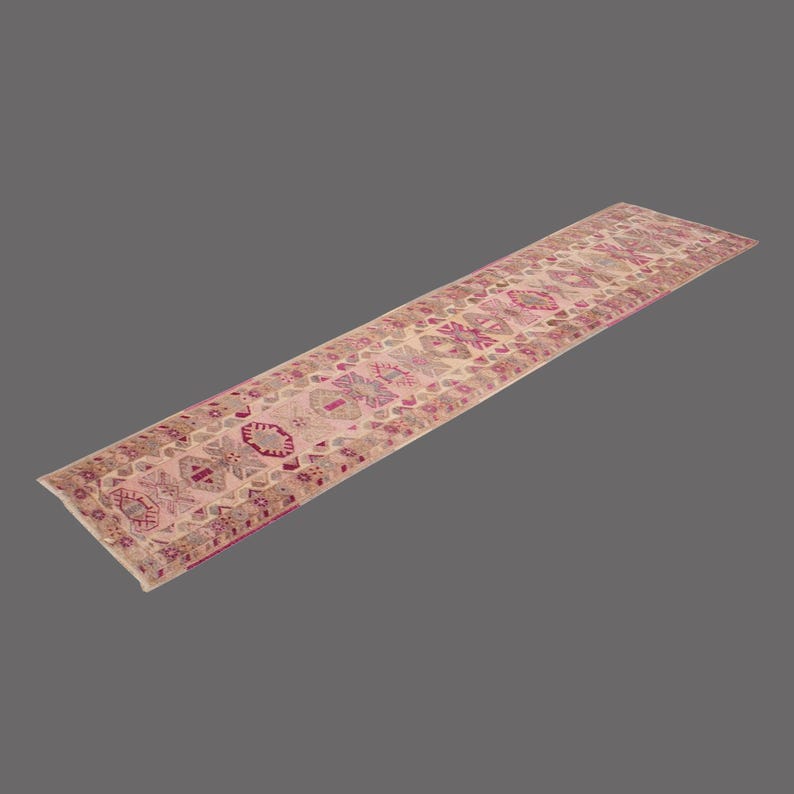 May include: A long, rectangular rug with a repeating floral pattern in shades of pink, beige, and burgundy. The rug has a distressed, vintage look, and is likely made of woven material. The rug is placed on a solid gray background.