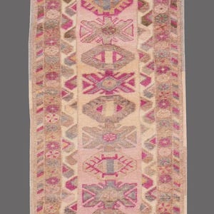 May include: A rectangular, patterned rug with a central design of repeating geometric shapes in pink, beige, and gray. The rug has a border with similar colors and patterns. The rug is likely made of woven fabric.