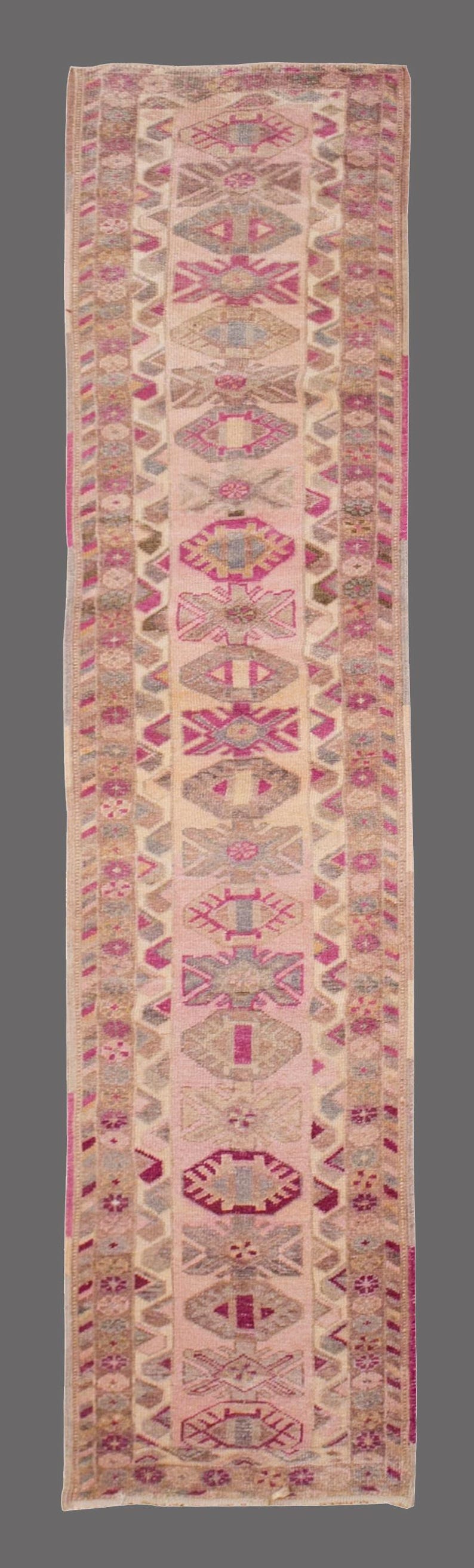 May include: A rectangular, patterned rug with a central design of repeating geometric shapes in pink, beige, and gray. The rug has a border with similar colors and patterns. The rug is likely made of woven fabric.