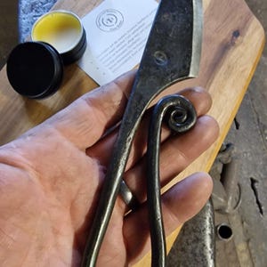 May include: Hand-forged, dark metal knife with a curved handle and a spiral design at the end. The blade is sharp and angled. A small tin of yellow balm and a wooden cutting board are also visible.