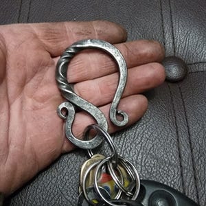 Può includere: Gancio portachiavi in metallo forgiato a mano con una sezione superiore attorcigliata e una serie di chiavi e un portachiavi per auto attaccati. Il gancio è color argento e ha un aspetto rustico e fatto a mano. Il testo "Warmmaiden and Berserk" è visibile.